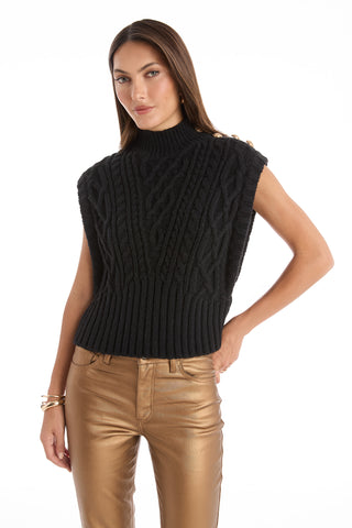 A woman wears the Raina Cable Knit Button Sweater in black with shiny metallic gold pants, standing with one hand on her hip against a white background.