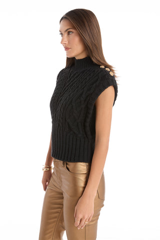 A woman in profile models the Raina Cable Knit Button Sweater in black—a sleeveless, mock turtleneck design with gold shoulder buttons—paired with tan leather pants against a plain white background.
