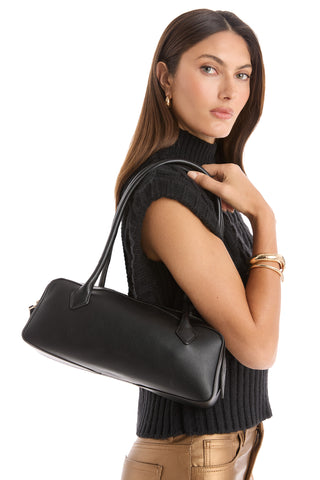 A woman with long brown hair, dressed in a sleeveless black sweater and gold pants, poses against a white background with the Reese Leather Bag - Black featuring gold hardware on her shoulder.