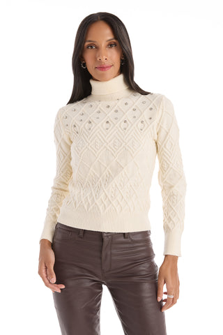 A woman with long dark hair wears the Cara Crystal Sweater - Pearl, a cream-colored turtleneck embellished with pearl accents, paired with brown leather pants, as she stands against a white background.