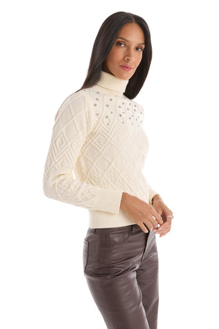 A woman with long dark hair models the Cara Crystal Sweater - Pearl, a cream-colored, pearl-embellished cable-knit turtleneck, paired with brown leather pants, standing against a white background.