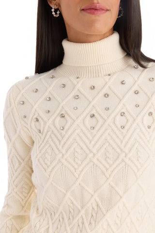 A woman wears the Cara Crystal Sweater - Pearl, a cream cable-knit turtleneck featuring a diamond pattern and jeweled accents on the shoulders and chest, with only her lower face, neck, and upper torso visible.