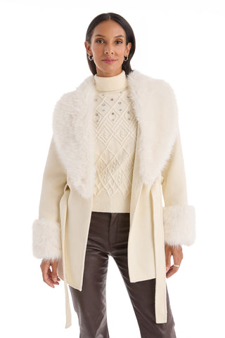 A woman wears the Isabel Fur Trim Coat in white, featuring a faux fur collar and cuffs, over a white diamond-patterned turtleneck and dark brown leather pants, standing against a white background.