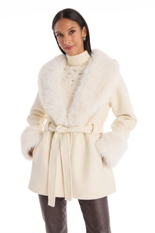 A woman wears the Isabel Fur Trim Coat in white, featuring a faux fur collar and cuffs with a belted waist, styled over a textured turtleneck sweater and dark pants, standing against a white background.