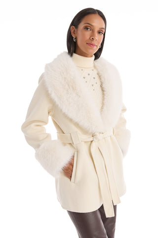 A woman wears the Isabel Fur Trim Coat in white, featuring a large faux fur collar and cuffs, a belted waist, and pockets. She pairs it with a light turtleneck and dark pants against a white background.