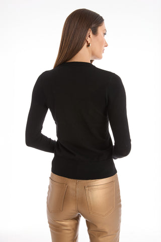 A woman with straight brown hair, wearing a fitted Marcela Knit Top - Black with gold buttons and metallic gold pants, stands facing away from the camera against a white background.