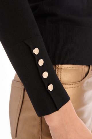 A close-up of a person's arm wearing the Marcela Knit Top - Black, a fitted sweater with four gold buttons at the cuff, paired with shiny gold-colored pants.
