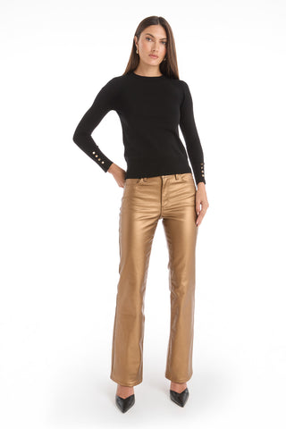 A woman faces forward against a plain white background wearing the Marcela Knit Top - Black with gold buttoned cuffs, shiny gold metallic pants, and black pointed-toe heels.