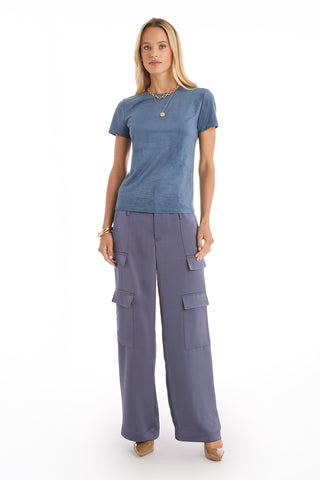 A woman faces forward against a white background, wearing the Amaya Suede T-Shirt in blue, wide-legged cargo pants in a matching shade, beige heels, and gold jewelry.