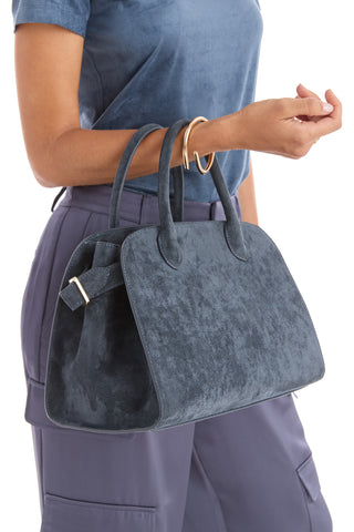 A person in a blue shirt and pants holds the Margo Suede Bag - Blue, featuring gold hardware and a classic silhouette, on their forearm against a white background.