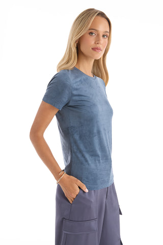 A woman with long blonde hair wears the Amaya Suede T-Shirt in Blue and dark cargo pants, standing with her hands in her pockets and facing slightly to the side against a plain white background.