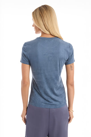 A woman with long blonde hair stands facing away, wearing the Amaya Suede T-Shirt in blue and loose blue-gray pants against a white background.