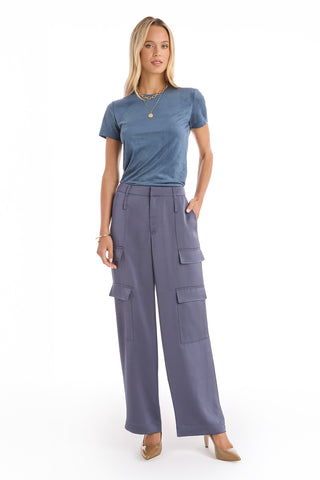 A woman with blond hair stands against a white background wearing navy Milan Satin Cargo Pants, a blue velvet t-shirt, tan heels, and layered gold necklaces. Her hands are in her pockets.