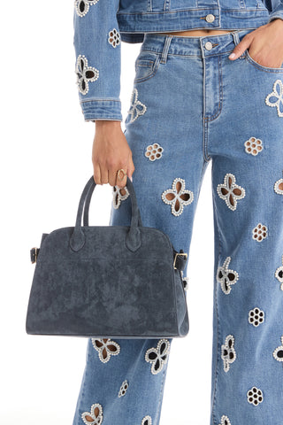 Wearing a blue denim jacket and jeans with white flower cutouts, a person holds the Margo Suede Bag - Blue by its handles. The focus is on the elegant bag, which features a detachable crossbody strap, and their lower torso.