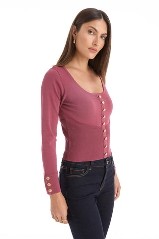 A woman with straight brown hair and gold earrings wears the Edie Knit Top in Sangria, featuring long sleeves and gold buttons, paired with dark blue jeans. She stands against a white background, looking at the camera neutrally.