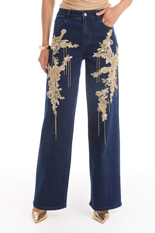 Wearing the Rocco Gold Embellished Jean in Indigo, featuring gold floral embroidery on the front thighs, paired with gold heels and a partially visible tan top.