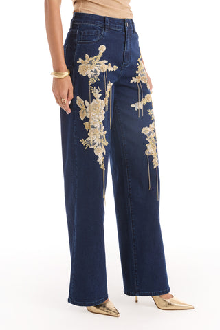 A person wearing the Rocco Gold Embellished Jean in indigo, a high-rise wide leg style with gold floral embroidery, paired with gold high-heeled shoes. Only their lower half is visible against a white background.