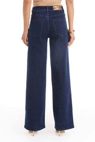 A person stands with their back turned, wearing the Rocco Gold Embellished Jean in indigo—a high-rise, wide-leg style with large back pockets and a leather waistband patch. Only the lower torso and legs are shown, paired with nude heels.