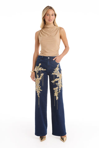 A woman stands wearing a sleeveless beige turtleneck top and the Rocco Gold Embellished Jean - Indigo, a high-rise, wide leg stretch denim accented with gold floral embroidery, styled with gold pointed-toe shoes.