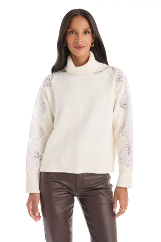A woman with long dark hair smiles softly as she faces forward, wearing the Camille Lace Turtleneck Sweater in Pearl and brown leather pants against a white background. The sweater features delicate lace sleeve detailing.