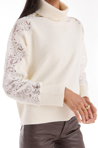 A person wearing the Camille Lace Turtleneck Sweater in Pearl, featuring delicate lace details on the shoulders and sleeves, styled with brown leather pants and gold rings.