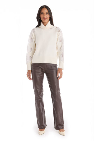 A woman faces forward against a white background, wearing the Camille Lace Turtleneck Sweater in Pearl, styled with brown leather pants and white pointed-toe heels.