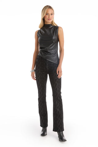A woman with straight blonde hair and a neutral expression poses against a white background wearing the Zaina Leather Top in black, styled sleeveless with an asymmetrical silhouette, paired with black textured pants, ankle boots, and silver bracelets.
