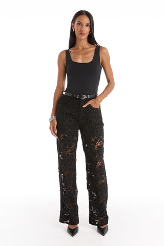 A woman stands facing forward, wearing a sleeveless black top, black belt, and Celeste High Rise Jean - Black. She has long, straight hair and is wearing black pointed heels.