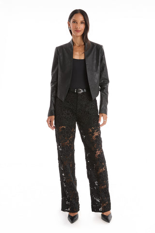 A woman stands facing forward in the Yasmin Leather Jacket - Black, with a black top, see-through black lace pants with a belt, and black pointed-toe shoes. The background is plain white.