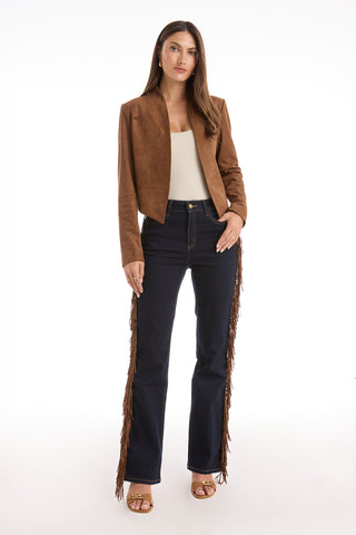 A woman faces forward in the Yasmin Suede Jacket - Cognac, featuring a draped front, over a white top, dark blue high-waisted fringe jeans, and tan heeled sandals against a plain white background.