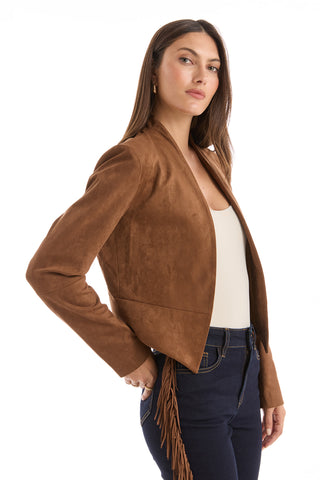 A woman stands confidently sideways against a white background, wearing the Yasmin Suede Jacket - Cognac featuring fringe and a draped front, paired with a cream top and dark blue jeans.