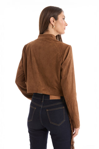A woman with long brown hair wears the Yasmin Suede Jacket - Cognac, a brown suede jacket with a draped front, and dark blue jeans. She stands with her back to the camera, looking to the side against a white background.