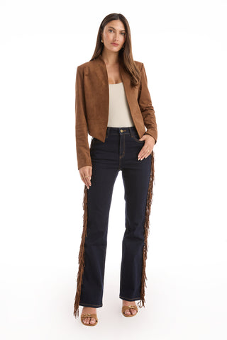 A woman with straight brown hair and a neutral expression faces forward, wearing a cream top, the Yasmin Suede Jacket - Cognac with a draped front, dark blue jeans with brown fringe on the sides, and brown heeled sandals.