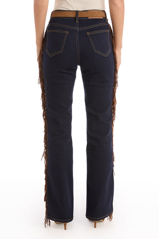 A person wearing the Ryder Fringe Jean - Indigo, a high-rise bootcut denim with brown fringe along the side seams and back pockets, stands with their back to the camera and pairs it with heeled shoes.