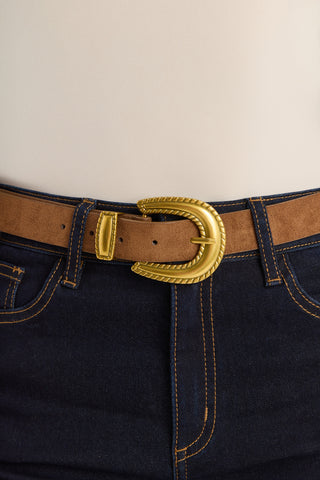 A close-up of a person wearing dark blue jeans and the Shelly Belt - Cognac with a large gold buckle, standing against a plain background.