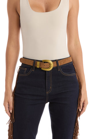 A person wears a beige sleeveless top with dark blue jeans and the Shelly Belt - Cognac, a western-inspired belt featuring a large gold cowboy buckle. The photo is cropped from shoulders to mid-thigh.