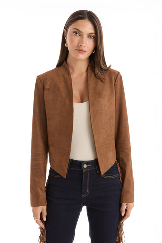 A woman with long brown hair stands against a white background, wearing the Yasmin Suede Jacket in Cognac over a white top and dark jeans.