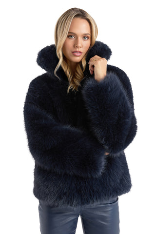 A woman with long blonde hair poses confidently against a plain white background, wearing the Dani Fur Coat - Navy, a dark blue cruelty-free faux fur coat with a high collar, matching cuffs, and dark pants.