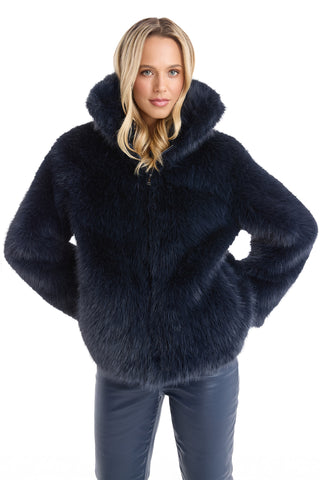 A woman with long blonde hair models the Dani Fur Coat - Navy, a stylish hooded faux fur coat, paired with matching pants and hands in pockets, against a white background—showcasing cruelty-free fashion.