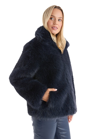 A woman with blonde hair models the Dani Fur Coat - Navy, a cruelty-free faux fur style, with one hand in her pocket, standing against a white background.