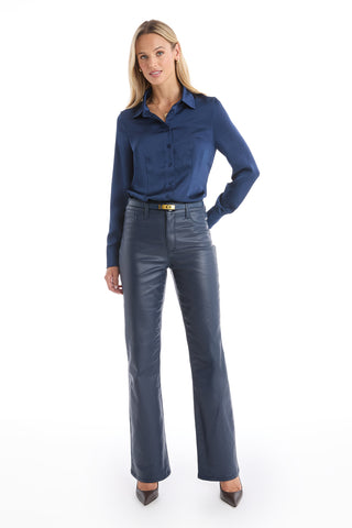 A woman stands against a white background, wearing the Aria Coated Flare Jean in Navy with a dark blue button-up shirt and pointed-toe heels. She has one hand in her pocket and smiles slightly.