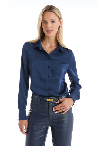 A woman with long blonde hair wears the Paulina Silk Blouse in navy, standing against a white background and looking at the camera with a slight smile, one hand resting on her hip.