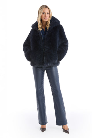 A woman with long blonde hair stands facing forward in the Dani Fur Coat - Navy, hands in her pockets. She pairs the cruelty-free faux fur coat with matching dark blue leather pants and pointed-toe heels against a white background.