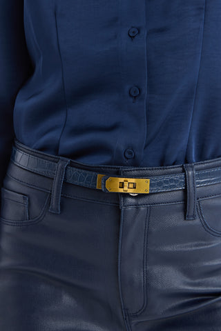 A person wearing a blue button-up shirt with blue textured pants, accessorized with the Nicola Belt - Navy, which features a croco-effect strap and a gold buckle.