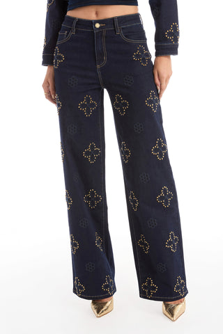 A person wearing the Kaia Studded High Rise Jean in indigo with gold studded floral stitching, a matching top, and metallic pointed-toe shoes poses against a white background.