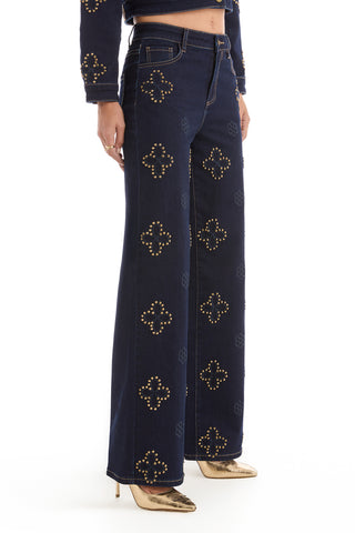 A person models the Kaia Studded High Rise Jean in indigo—high-waisted, wide-leg denim adorned with gold geometric studs—paired with gold pointed-toe heels. The image is cropped at the waist and ankles.