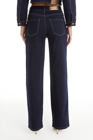 Person facing away wears Kaia Studded High Rise Jean - Indigo: dark blue, high-rise, straight-leg jeans with visible stitching and back pockets. Paired with a matching denim jacket and tan heeled shoes.