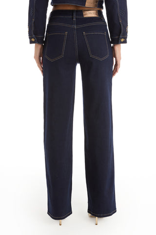 A person stands from the back against a white background, wearing Kaia Studded High Rise Jean in indigo with a matching jacket and light-colored high heels.