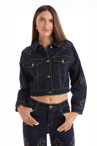A woman models the Kaia Studded Jacket - Indigo, a dark blue denim jacket with gold studs and floral stitching, paired with matching high-waisted jeans. She stands confidently with one hand in her pocket against a white background.