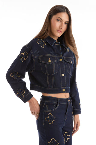 A woman with long brown hair wears the Kaia Studded Jacket - Indigo, featuring gold studs in floral and diamond patterns, paired with matching jeans. She poses with one hand on her hip against a white background.